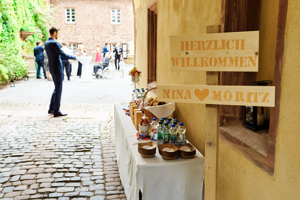 Onelovephoto Ein Holzschild mit der Aufschrift "Herzlich Willkommen Nina ♥ Moritz" steht neben einem Tisch mit Getränken und Snacks. Im Hintergrund versammeln sich Menschen und unterhalten sich in einem Hof mit Kopfsteinpflaster und warmen gelben und roten Backsteinwänden. Hochzeitsfotografie in Mannheim und Heidelberg