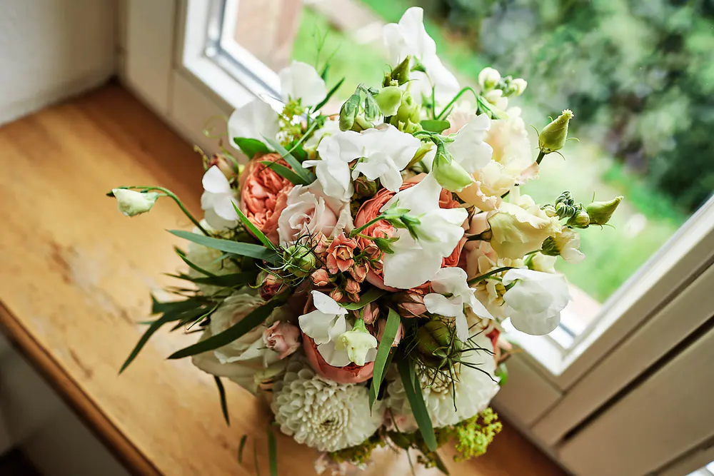 Onelovephoto Ein Strauß weißer, pfirsichfarbener und rosafarbener Blumen mit grünem Laub steht auf einer hölzernen Fensterbank. Das Sonnenlicht fällt durch das Fenster, und draußen ist ein üppiger grüner Garten zu sehen. Hochzeitsfotografie in Mannheim und Heidelberg