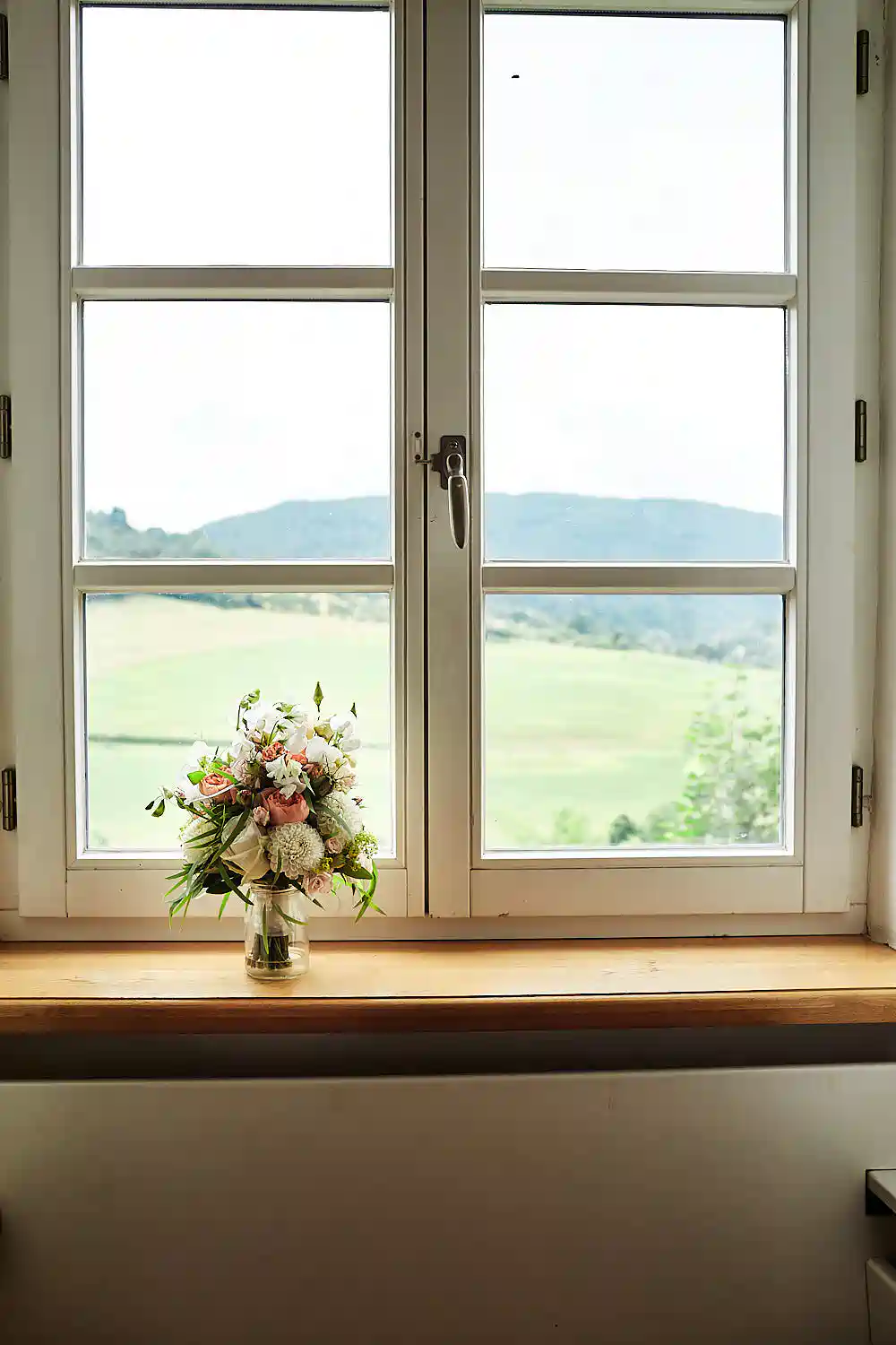 Onelovephoto Eine Vase mit Blumen steht auf einer hölzernen Fensterbank vor einem großen Fenster mit Blick auf grüne Felder und ferne Hügel. Hochzeitsfotografie in Mannheim und Heidelberg