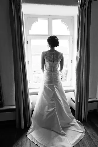 Eine Braut in einem weißen Kleid steht mit dem Rücken zur Kamera und blickt aus einem hellen, von Vorhängen umrahmten Fenster in einem sanft beleuchteten Raum - so wird der Zauber von Heiraten auf der Mittelburg in Neckarsteinach perfekt eingefangen.