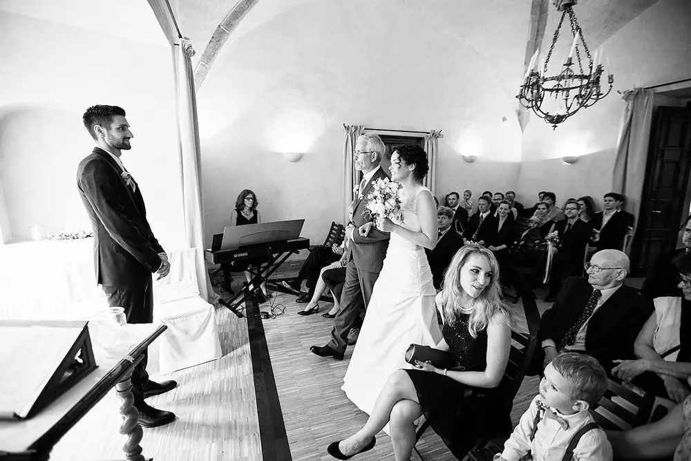 Onelovephoto Eine Braut schreitet mit einem älteren Mann in einer kleinen, überdachten Hochzeitszeremonie zum Altar, während der Bräutigam am Eingang wartet. Die Gäste sitzen und schauen zu, während im Hintergrund ein Klavierspieler spielt. Die Szene ist in schwarz-weiß gehalten. Hochzeitsfotografie in Mannheim und Heidelberg
