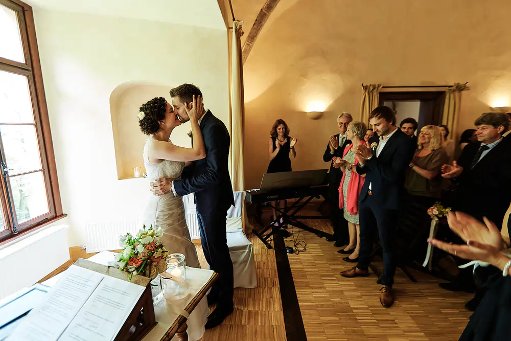 Onelovephoto Eine Braut und ein Bräutigam küssen sich während ihrer Hochzeitszeremonie in einem warm beleuchteten Raum, während die Gäste um sie herum stehen und applaudieren. Ein Blumenstrauß und ein offenes Buch liegen auf einem Tisch in der Nähe. Hochzeitsfotografie in Mannheim und Heidelberg