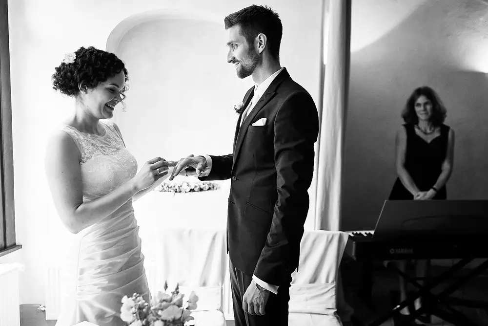 Onelovephoto Eine Braut steckt dem Bräutigam während einer Hochzeitszeremonie einen Ring an den Finger, beide lächeln. Eine Frau steht im Hintergrund neben einer Tastatur und beobachtet das Paar. Die Szene ist in schwarz-weiß gehalten. Hochzeitsfotografie in Mannheim und Heidelberg