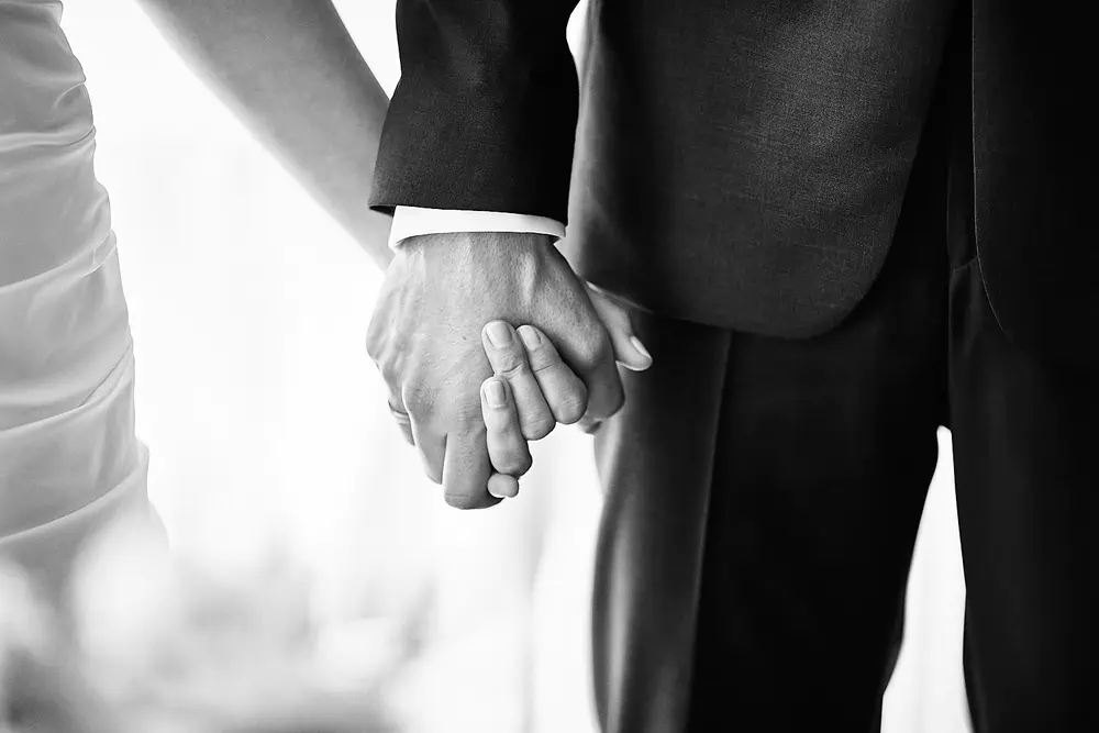 Onelovephoto Schwarz-Weiß-Foto in Nahaufnahme eines Paares, das sich an den Händen hält und formell gekleidet ist; eine Person trägt einen Anzug, die andere ein Kleid, was auf eine Hochzeit oder einen besonderen Anlass hindeutet. Hochzeitsfotografie in Mannheim und Heidelberg