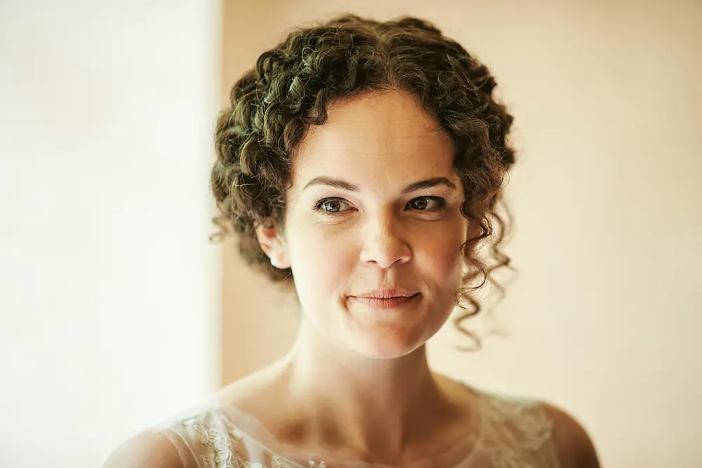 Onelovephoto Eine Frau mit lockigem braunem Haar in einer Hochsteckfrisur lächelt sanft. Sie trägt ein durchsichtiges, spitzenbesetztes Oberteil und wird von einem weichen, natürlichen Licht vor einem beigen Hintergrund beleuchtet. Hochzeitsfotografie in Mannheim und Heidelberg