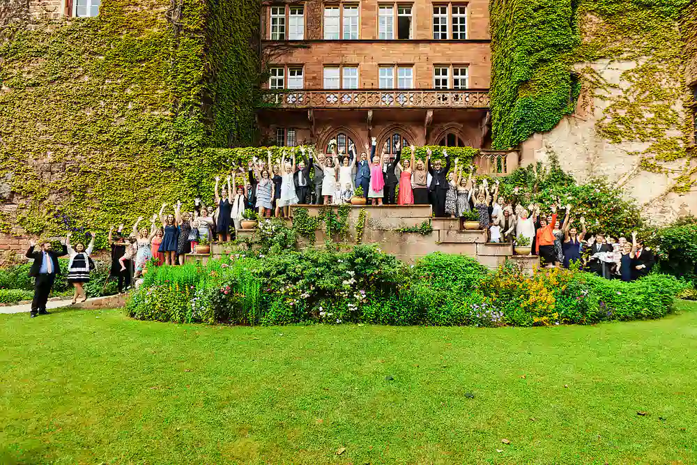Onelovephoto Eine große Gruppe förmlich gekleideter Menschen steht auf gestuften Steinstufen vor einem mit Efeu bewachsenen historischen Gebäude und hebt feierlich die Arme. Im Vordergrund sind üppiges grünes Gras und blühende Pflanzen zu sehen. Hochzeitsfotografie in Mannheim und Heidelberg