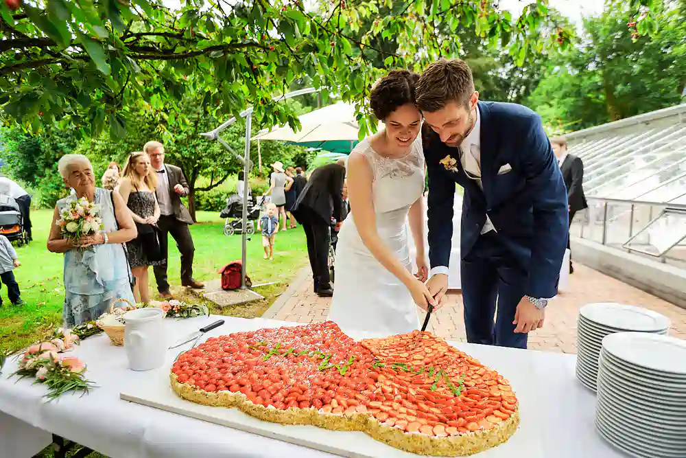 Onelovephoto Braut und Bräutigam lächeln, als sie an ihrem Hochzeitstag im Freien eine große, herzförmige Erdbeertorte anschneiden, während die Gäste zuschauen und das Grün im Hintergrund zu sehen ist. Hochzeitsfotografie in Mannheim und Heidelberg