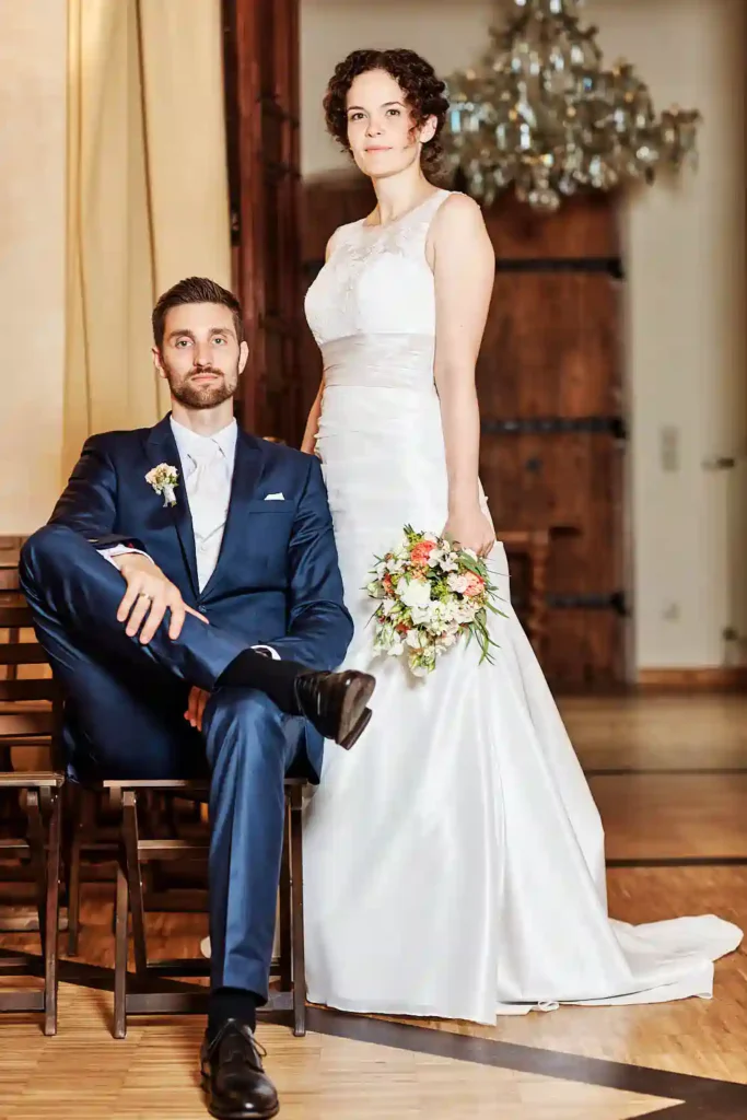 Eine Braut in einem weißen Kleid mit einem Blumenstrauß in der Hand steht neben einem sitzenden Bräutigam in einem marineblauen Anzug; beide schauen in einer eleganten Inneneinrichtung mit Holz- und Kronleuchterdetails in die Kamera - perfekt für heiraten auf der Mittelburg in Neckarsteinach. Onelovephoto - Hochzeitsfotografie in Mannheim und Heidelberg
