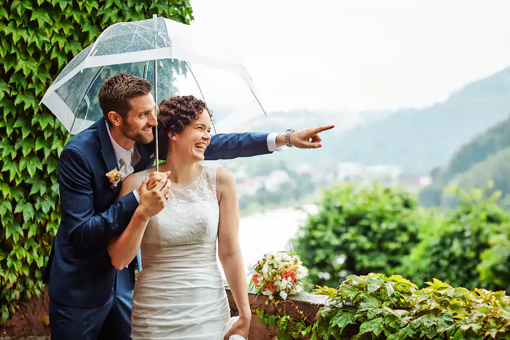 Eine lächelnde Braut und ein lächelnder Bräutigam stehen im Freien unter einem klaren Regenschirm. Der Bräutigam umarmt die Braut von hinten und gestikuliert aufgeregt in die Ferne - perfekte Momente für heiraten auf der Mittelburg in Neckarsteinach mit üppigem Grün und Hügeln ringsum. Onelovephoto - Hochzeitsfotografie in Mannheim und Heidelberg