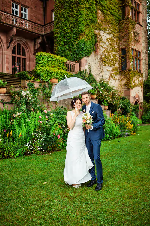 Eine Braut und ein Bräutigam stehen bei ihrer Hochzeit auf der Mittelburg Neckarsteinach zusammen auf grünem Gras vor einem efeubewachsenen Gebäude und teilen sich einen durchsichtigen Regenschirm. Die Braut ist weiß gekleidet, der Bräutigam trägt einen blauen Anzug. Üppige Gartenblumen umgeben sie.