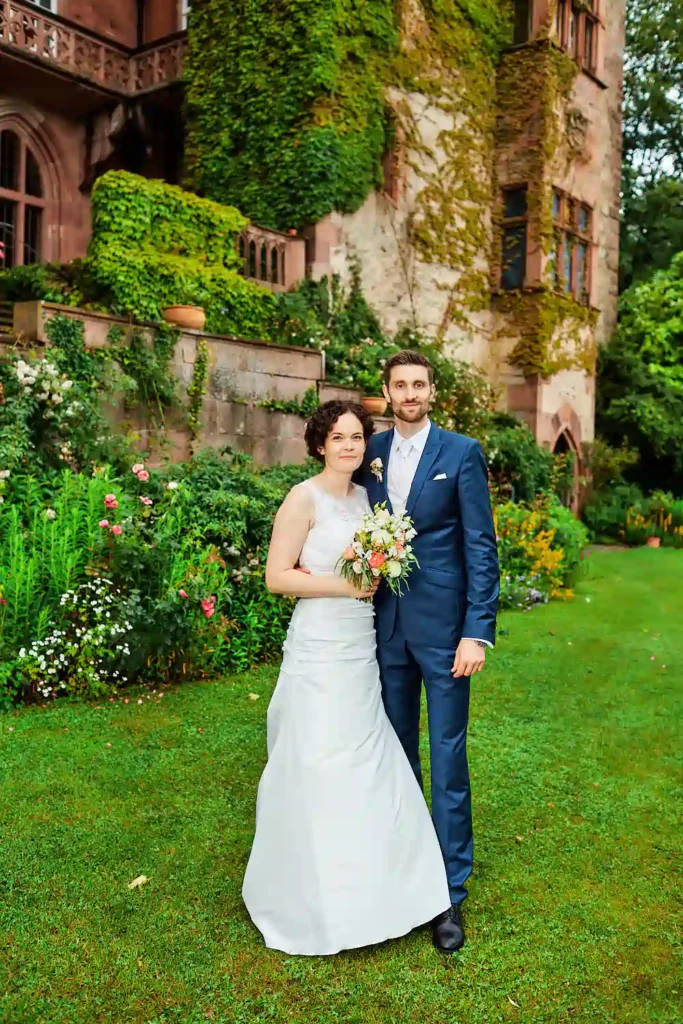 Eine Braut im weißen Kleid und ein Bräutigam im blauen Anzug stehen zusammen auf einer grünen Wiese, umgeben von Blumen, mit einem mit Weinreben bewachsenen historischen Gebäude im Hintergrund - perfekt für heiraten auf der Mittelburg in Neckarsteinach. Onelovephoto - Hochzeitsfotografie in Mannheim und Heidelberg