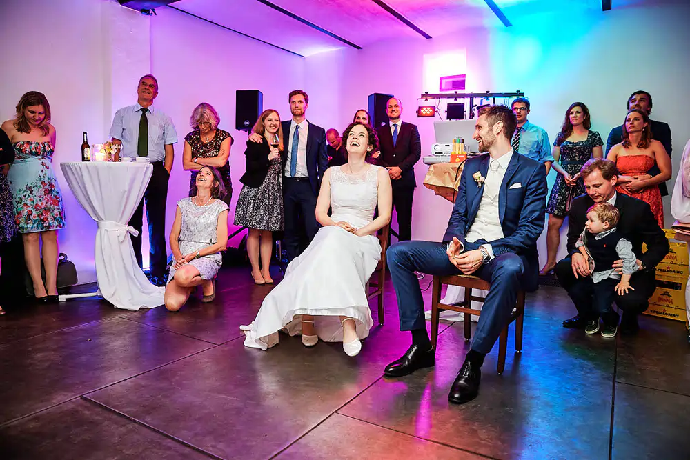 Braut und Bräutigam sitzen lächelnd in der Mitte eines hell erleuchteten Raumes während ihrer Heiraten auf der Mittelburg in Neckarsteinach, umgeben von förmlich gekleideten Freunden und Familienmitgliedern, die gemeinsam einen festlichen Moment genießen. Onelovephoto - Hochzeitsfotografie in Mannheim und Heidelberg