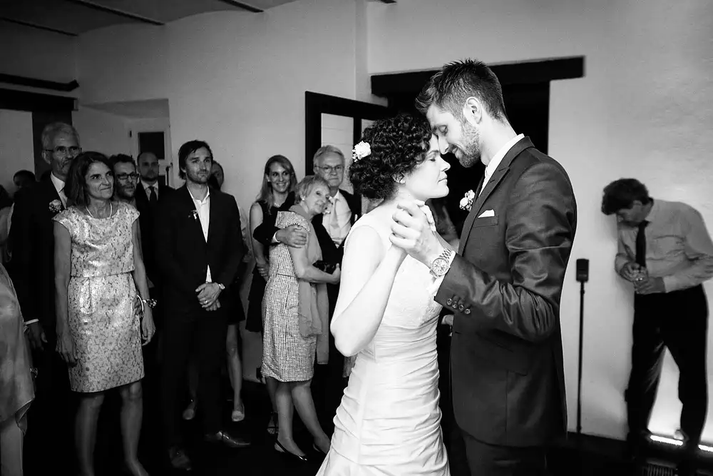 Braut und Bräutigam tanzen eng beieinander, berühren sich an den Stirnen und halten sich an den Händen, während die Hochzeitsgäste im Hintergrund lächelnd zusehen. Dieser intime Moment wurde in Schwarz-Weiß aufgenommen und entfaltet sich während der Heiraten auf der Mittelburg in Neckarsteinach. Onelovephoto - Hochzeitsfotografie in Mannheim und Heidelberg