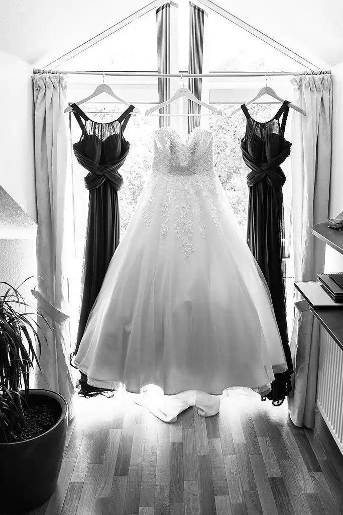 Onelovephoto Ein weißes trägerloses Hochzeitskleid und zwei dunkle Brautjungfernkleider hängen auf Kleiderbügeln vor einem hellen Fenster mit Vorhängen, das von natürlichem Licht in einem Raum mit Holzfußboden beleuchtet wird. Hochzeitsfotografie in Mannheim und Heidelberg