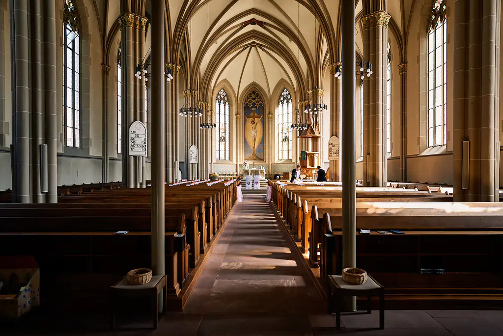 Das Innere einer großen Kirche mit hölzernen Kirchenbänken, hohen Gewölbedecken, Buntglasfenstern und einem Mittelgang, der zu einem verzierten Altar mit einem Kruzifix führt - perfekt für eine Hochzeit in der Stadtkirche Walldorf, wenn das Sonnenlicht durch die Fenster strömt. Onelovephoto - Hochzeitsfotografie in Mannheim und Heidelberg