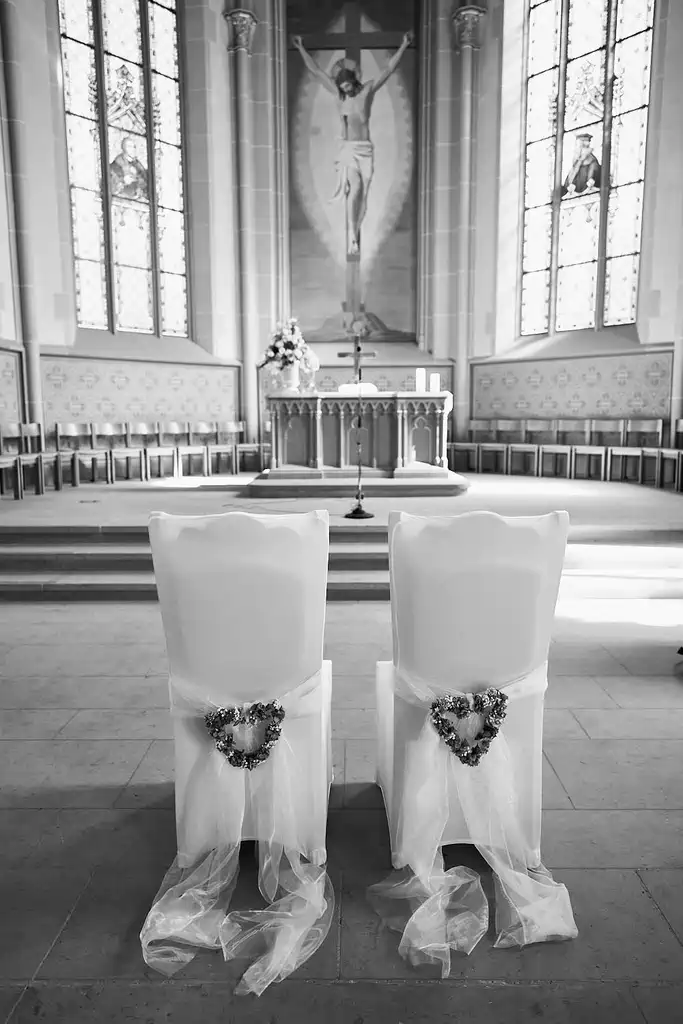 Zwei weiße Stühle, geschmückt mit herzförmigen Kränzen und durchsichtigem Stoff, stehen vor dem Altar in der Kirche, bereit für eine Hochzeit in der Stadtkirche Walldorf. Buntglasfenster und ein großes Kruzifix bilden eine schöne Hochzeitsszene. Onelovephoto - Hochzeitsfotografie in Mannheim und Heidelberg