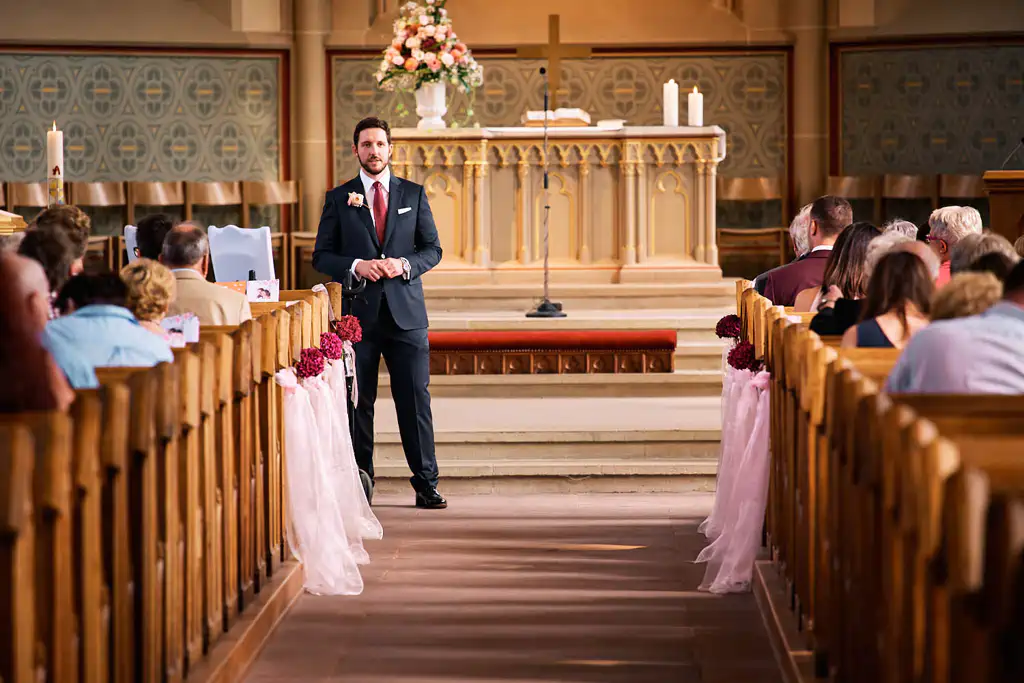 Onelovephoto Ein Mann im Anzug steht am Anfang eines mit rosa Bändern geschmückten Kirchenschiffs, während die Gäste in den Kirchenbänken auf den Beginn einer Zeremonie warten. Im Hintergrund ist ein Altar mit Kerzen und Blumen zu sehen. Hochzeitsfotografie in Mannheim und Heidelberg