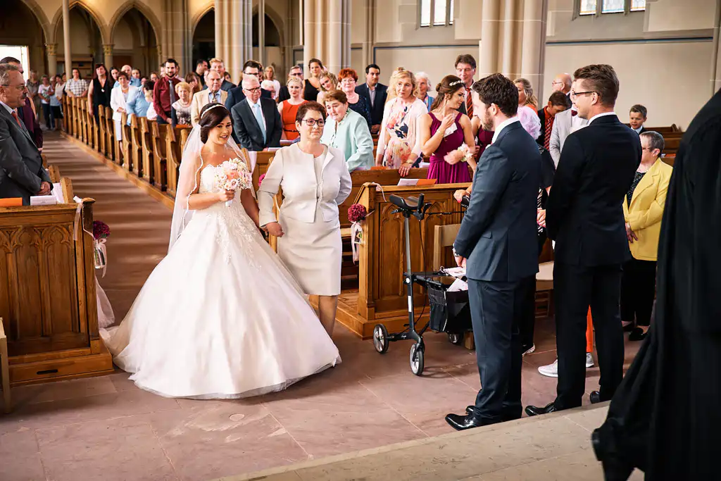 Onelovephoto Eine Braut in einem weißen Kleid schreitet den Gang hinunter, an ihrer Seite eine ältere Frau in einem weißen Outfit. Die Gäste stehen und beobachten, wie sie sich zwei Männern nähern, die vorne in einer Kirche mit hohen Bögen warten. Hochzeitsfotografie in Mannheim und Heidelberg