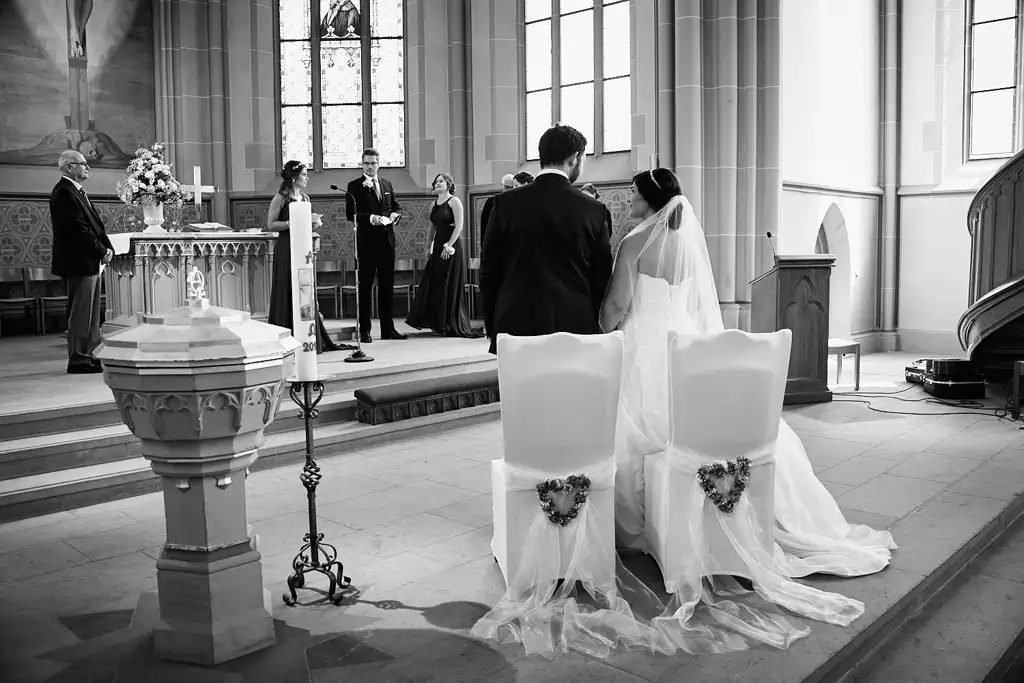 Braut und Bräutigam knien während ihrer Hochzeit in der Stadtkirche Walldorf mit dem Rücken zur Kamera vor dem Altar in einer Kirche. Die Gäste und der Trauzeuge sind zu sehen, gebadet in natürlichem Licht aus hohen Fenstern. Onelovephoto - Hochzeitsfotografie in Mannheim und Heidelberg