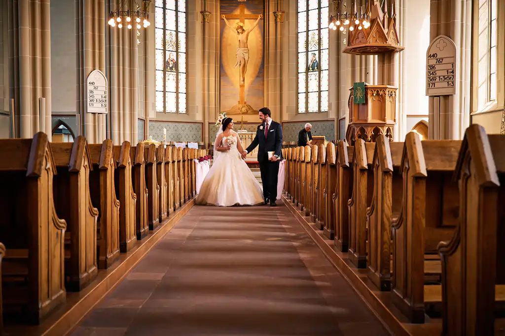 Braut und Bräutigam schreiten Hand in Hand den Gang der prächtigen Stadtkirche Walldorf entlang. Die hohen Glasfenster, die hölzernen Kirchenbänke und das große Kruzifix machen ihre Hochzeit in der Stadtkirche Walldorf unvergesslich. Onelovephoto - Hochzeitsfotografie in Mannheim und Heidelberg
