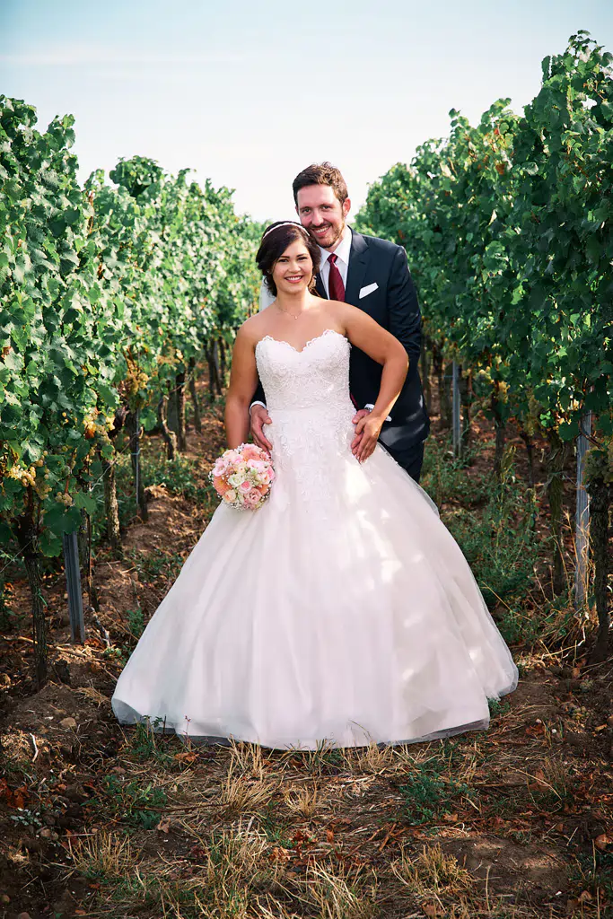 Onelovephoto Eine Braut in einem weißen Kleid mit einem Blumenstrauß in der Hand steht vor einem Bräutigam in einem dunklen Anzug, beide lächeln, umgeben von grünen Weinstöcken in einem Weinberg im Freien. Hochzeitsfotografie in Mannheim und Heidelberg
