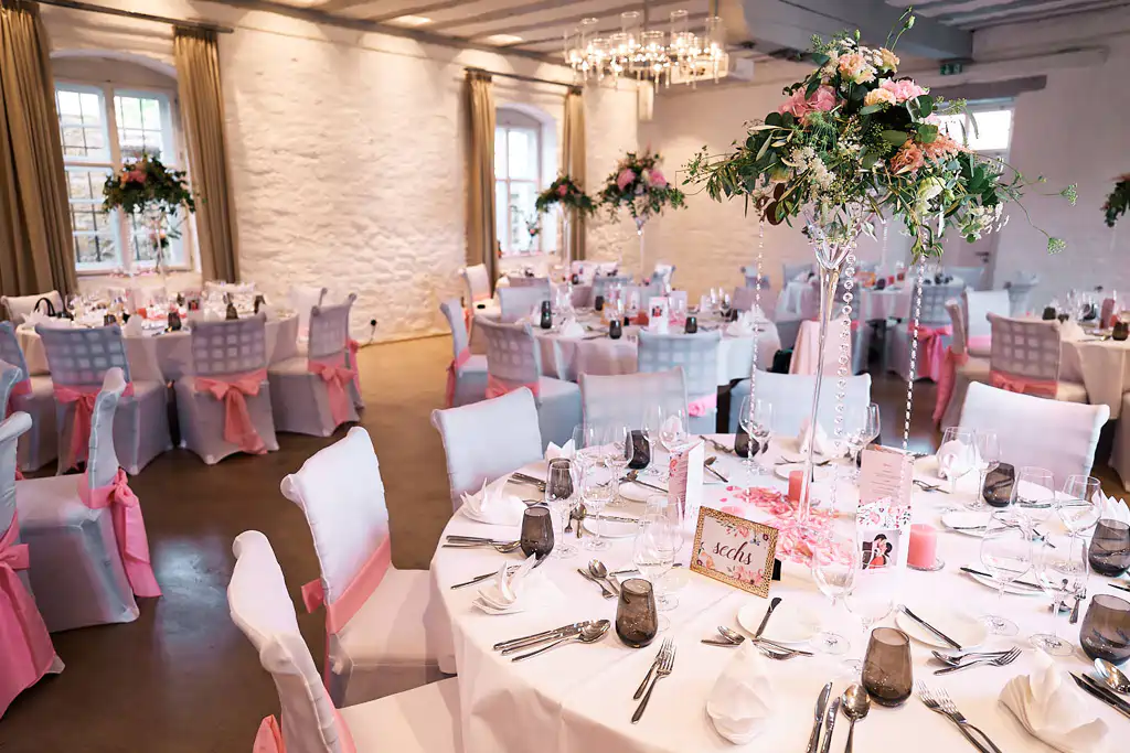Eleganter Bankettsaal für eine Veranstaltung mit runden Tischen mit weißen Tüchern, rosa Dekoration und Blumenschmuck - perfekt für eine Hochzeit im Ketschauer Hof Deidesheim. Helles und luftiges Ambiente mit Stühlen mit weißen Hussen und rosa Schärpen. Onelovephoto - Hochzeitsfotografie in Mannheim und Heidelberg