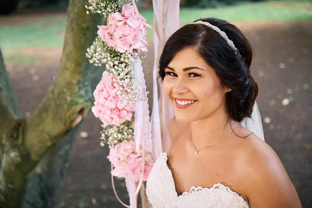 Eine Braut in weißem Kleid und Stirnband lächelt im Freien neben einem mit rosa Blumen und weißen Bändern geschmückten Baum und fängt damit den Zauber einer Hochzeit im Ketschauer Hof Deidesheim ein. Onelovephoto - Hochzeitsfotografie in Mannheim und Heidelberg
