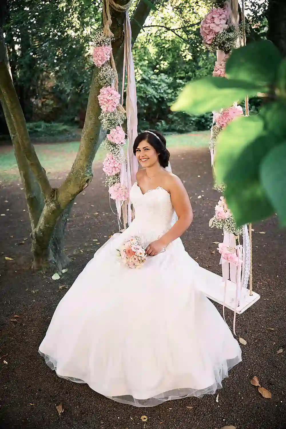 Onelovephoto Eine Braut in einem weißen Hochzeitskleid sitzt auf einer mit rosa Blumen geschmückten Schaukel, lächelt und hält einen Blumenstrauß in der Hand, umgeben von Bäumen in einer Gartenlandschaft. Hochzeitsfotografie in Mannheim und Heidelberg