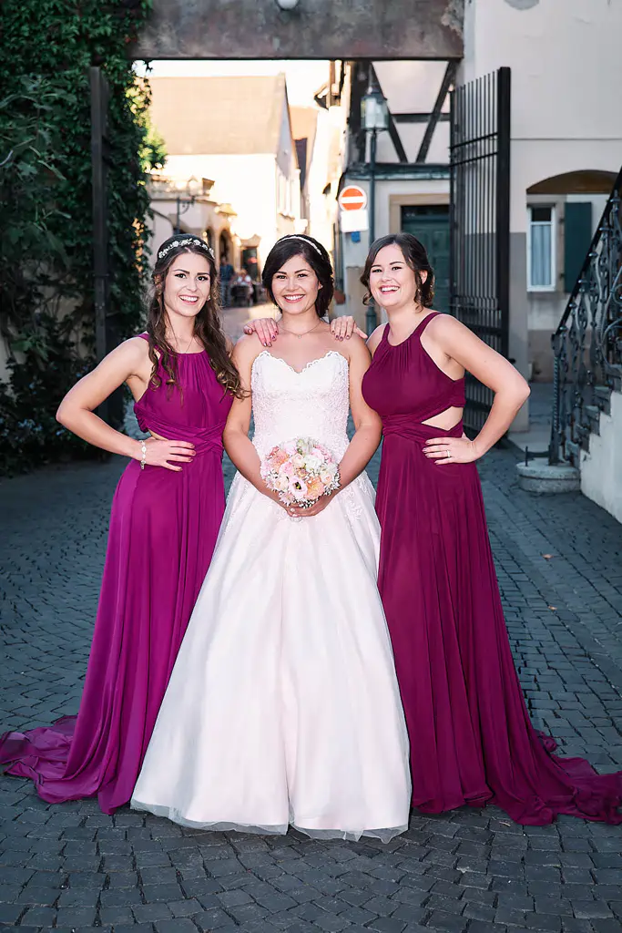 Onelovephoto Drei Frauen stehen lächelnd beieinander, die Braut in einem weißen Kleid mit einem Blumenstrauß in der Hand, flankiert von zwei Brautjungfern in passenden dunkelvioletten Kleidern, im Freien auf einer Straße mit Kopfsteinpflaster. Hochzeitsfotografie in Mannheim und Heidelberg