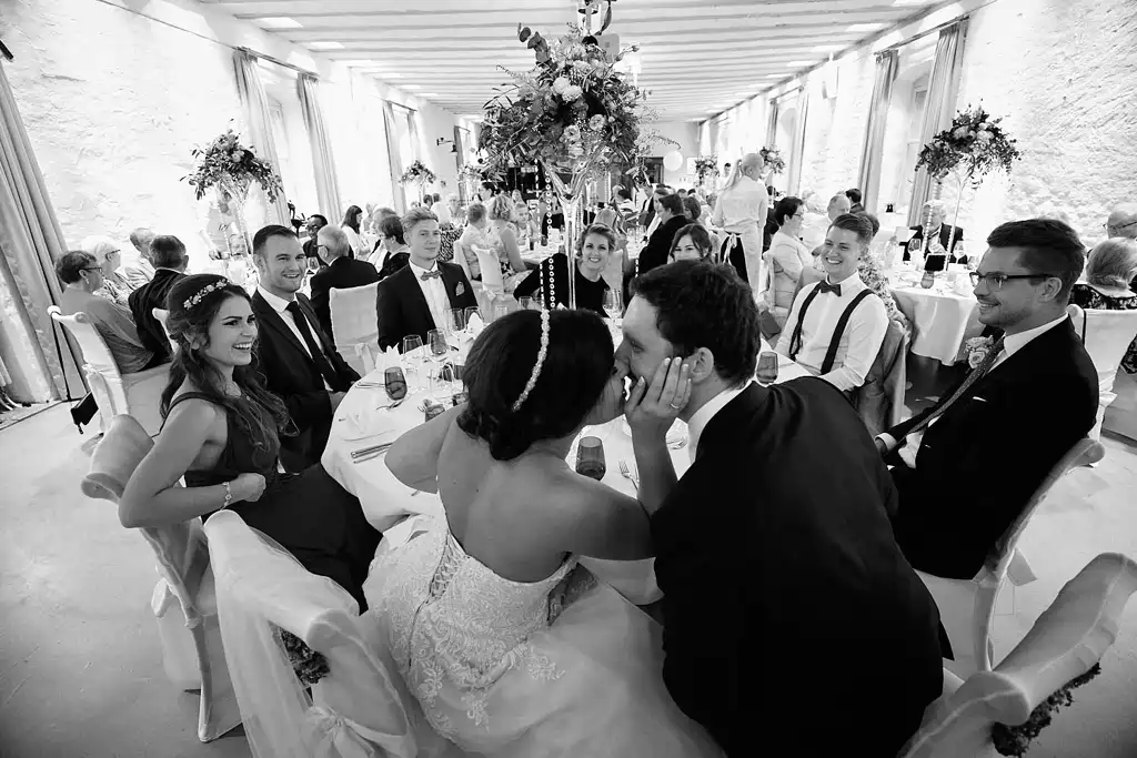 Onelovephoto Braut und Bräutigam küssen sich an einem runden Tisch, umgeben von lächelnden Gästen in formeller Kleidung bei einem Hochzeitsempfang in einem hellen, eleganten und mit Blumen geschmückten Saal. Hochzeitsfotografie in Mannheim und Heidelberg