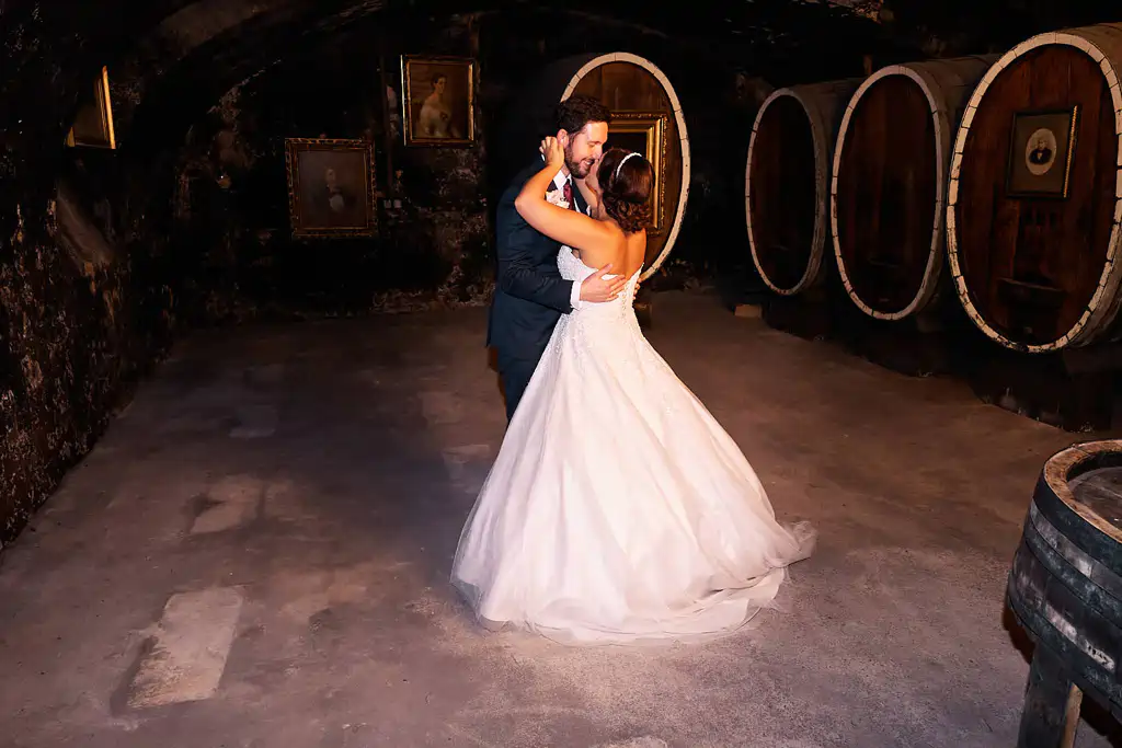 Onelovephoto Eine Braut und ein Bräutigam tanzen ihren ersten Tanz in einem schwach beleuchteten Keller mit großen Fässern und gerahmten Porträts an den Wänden. Die Braut trägt ein weißes Kleid, der Bräutigam einen dunklen Anzug. Hochzeitsfotografie in Mannheim und Heidelberg