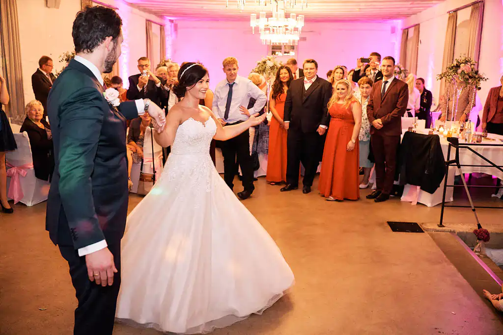 Onelovephoto Eine Braut und ein Bräutigam tanzen zusammen in einer hellen, dekorierten Empfangshalle, umgeben von Gästen, die zusehen und lächeln. Die Braut trägt ein weißes Kleid und der Bräutigam einen dunklen Anzug. Der Raum ist mit warmer Beleuchtung und Blumen festlich geschmückt. Hochzeitsfotografie in Mannheim und Heidelberg