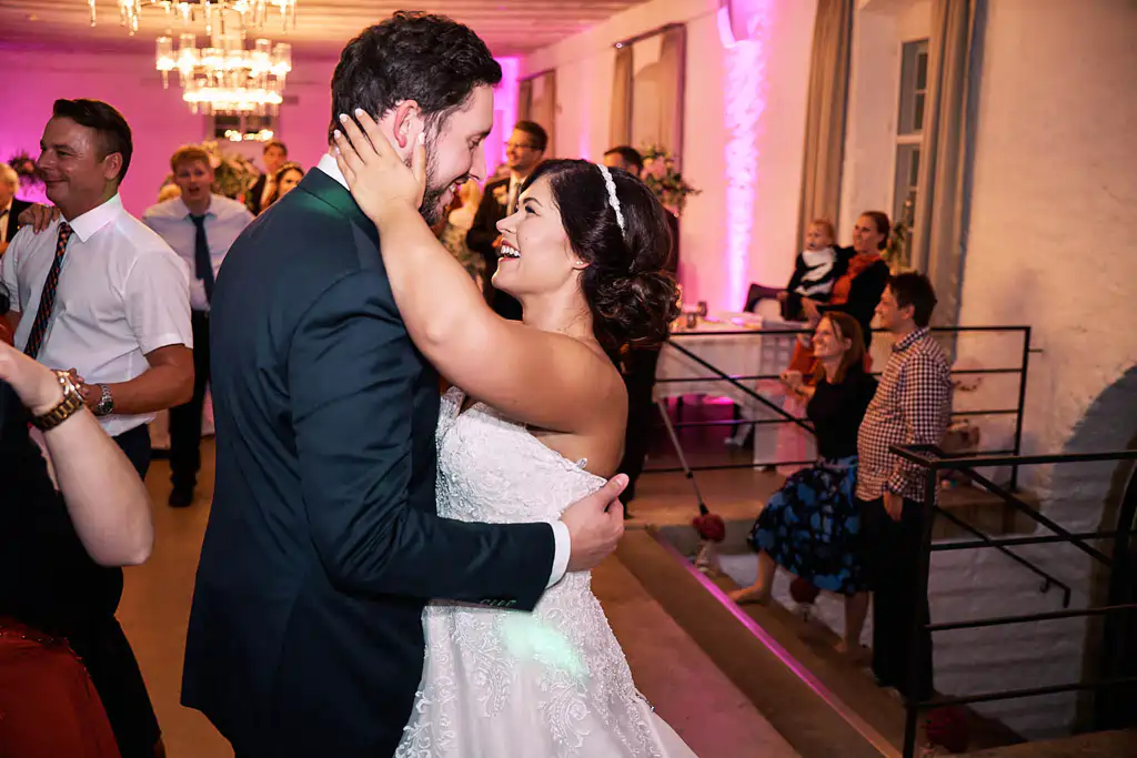 Onelovephoto Braut und Bräutigam lächeln und umarmen sich beim Tanzen auf ihrer Hochzeitsfeier, umgeben von jubelnden Gästen in einem warm beleuchteten Raum mit rosafarbener Deckenbeleuchtung. Hochzeitsfotografie in Mannheim und Heidelberg