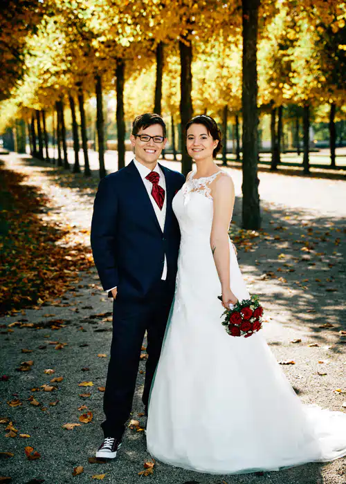 Onelovephoto Eine lächelnde Braut in einem weißen Kleid mit einem roten Blumenstrauß steht neben einem Bräutigam in einem marineblauen Anzug und Turnschuhen. Sie posieren auf einem von Herbstblättern gesäumten Weg unter einem Baldachin aus goldenem Laub. Hochzeitsfotografie in Mannheim und Heidelberg Onelovephoto Eine lächelnde Braut in einem weißen Kleid mit einem roten Blumenstrauß steht neben einem Bräutigam in einem marineblauen Anzug und Turnschuhen. Sie posieren auf einem von Herbstblättern gesäumten Weg unter einem Baldachin aus goldenem Laub. Hochzeitsfotografie in Mannheim und Heidelberg