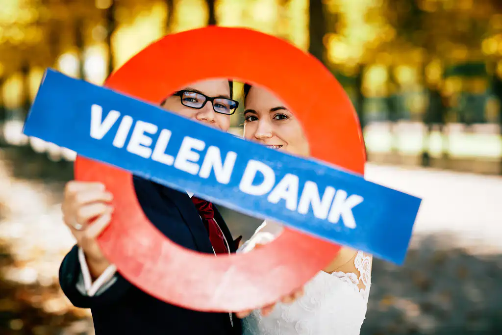 Onelovephoto Ein lächelndes Paar in Hochzeitskleidung hält einen runden roten Rahmen mit einem blauen Schild, auf dem "VIELEN DANK" steht; im Hintergrund sind Bäume und Sonnenlicht zu sehen. Hochzeitsfotografie in Mannheim und Heidelberg