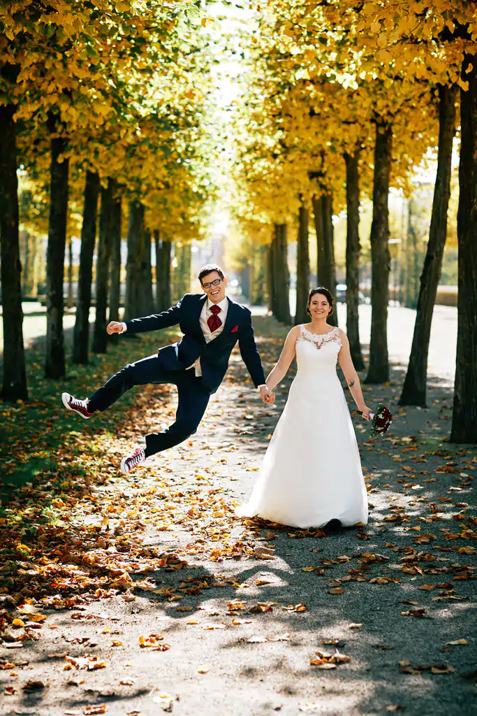 Onelovephoto Eine Braut und ein Bräutigam stehen auf einem von Bäumen gesäumten Weg mit Herbstlaub. Der Bräutigam, der einen Anzug und rote Turnschuhe trägt, springt spielerisch umher und hält die Hand der Braut. Die Braut trägt ein weißes Kleid, lächelt und hält einen Blumenstrauß. Hochzeitsfotografie in Mannheim und Heidelberg