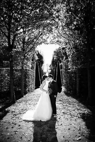 Eine Braut und ein Bräutigam stehen eng beieinander Onelovephoto Eine Braut und ein Bräutigam stehen eng beieinander auf einem sonnenbeschienenen, von Bäumen gesäumten Weg in einem Garten und küssen sich. Das Schwarz-Weiß-Foto fängt ihren romantischen Moment ein, während die Blätter auf dem Boden verstreut sind. Hochzeitsfotografie in Mannheim und Heidelberg