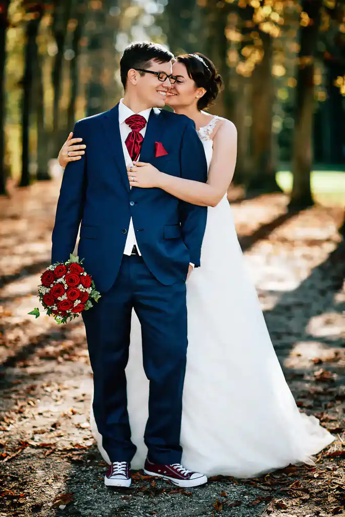Eine Braut in einem weißen Kleid umarmt und küsst einen lächelnden Bräutigam in einem blauen Anzug, der rote Blumen in der Hand hält. Als Hochzeitsfotograf in Schwetzingen habe ich diesen romantischen Moment auf einem sonnendurchfluteten, von Bäumen gesäumten Weg im Freien eingefangen. Onelovephoto - Hochzeitsfotografie in Mannheim und Heidelberg