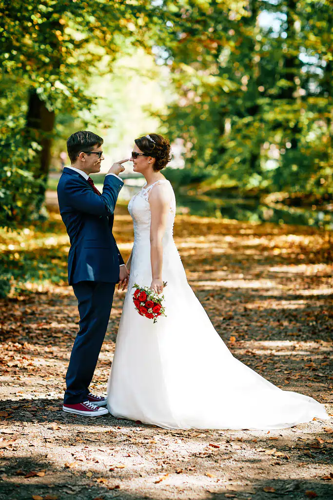 Onelovephoto Eine Braut in einem weißen Kleid mit einem Blumenstrauß steht einem Bräutigam in einem marineblauen Anzug und roten Turnschuhen gegenüber. Sie stehen auf einem sonnenbeschienenen, laubbedeckten Weg in einer grünen, bewaldeten Gegend und lächeln sich an. Hochzeitsfotografie in Mannheim und Heidelberg