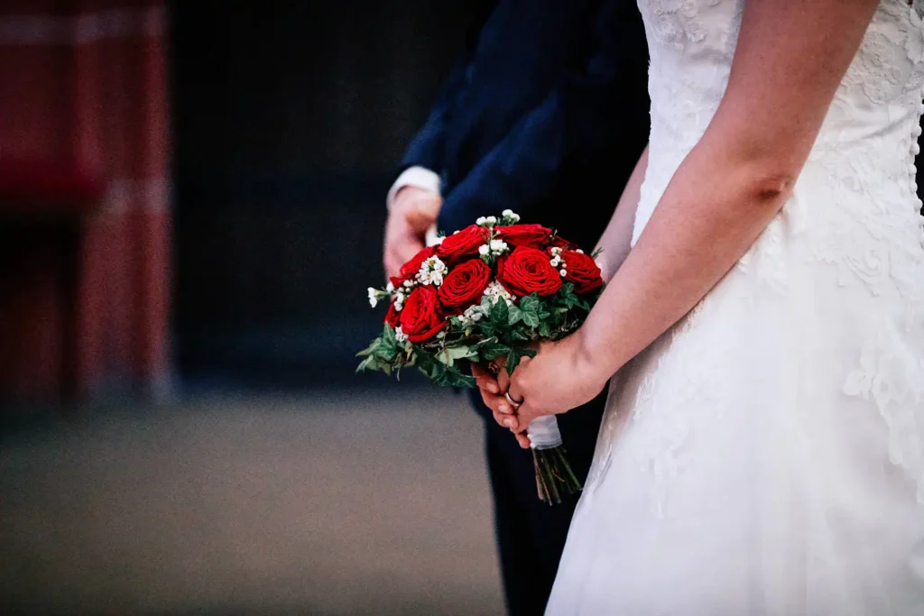 Onelovephoto Eine Braut in einem weißen Spitzenkleid hält einen Strauß roter Rosen und Grünzeug in der Hand und steht neben einer Person in einem dunklen Anzug; beide sind teilweise sichtbar, wobei der Schwerpunkt auf dem Strauß und ihren Händen liegt. Hochzeitsfotografie in Mannheim und Heidelberg