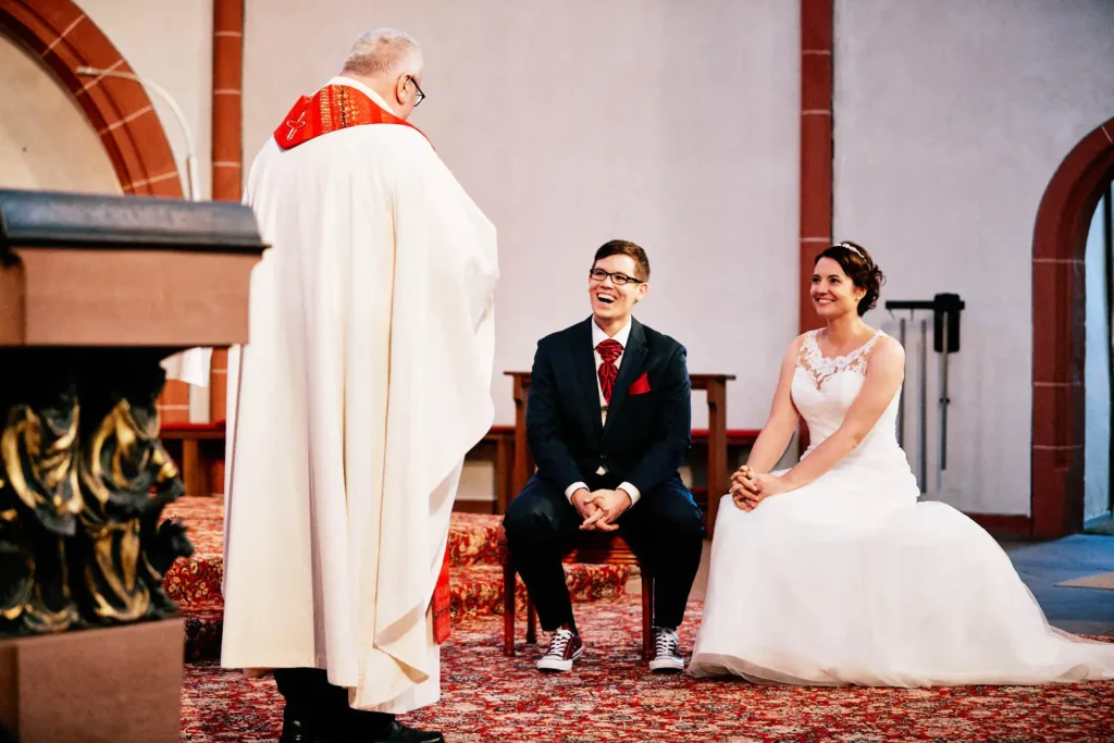 Onelovephoto Eine Braut und ein Bräutigam sitzen lächelnd und lachend während ihrer Hochzeitszeremonie vor einem Zeremonienmeister in weißem Gewand in einer Kirche mit rotem Teppich und Steinbögen. Hochzeitsfotografie in Mannheim und Heidelberg