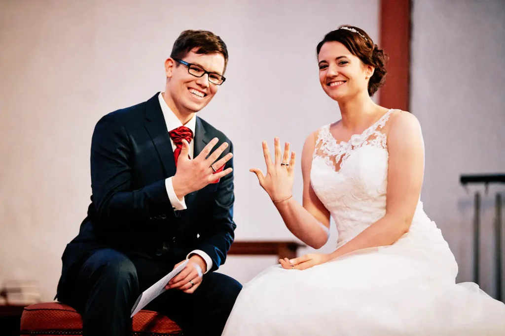 Onelovephoto Ein frisch verheiratetes Paar in Hochzeitskleidung sitzt Seite an Seite, lächelt und zeigt ihre Eheringe an der linken Hand. Der Bräutigam trägt einen dunklen Anzug mit einer roten Fliege, die Braut ein weißes Spitzenkleid. Hochzeitsfotografie in Mannheim und Heidelberg