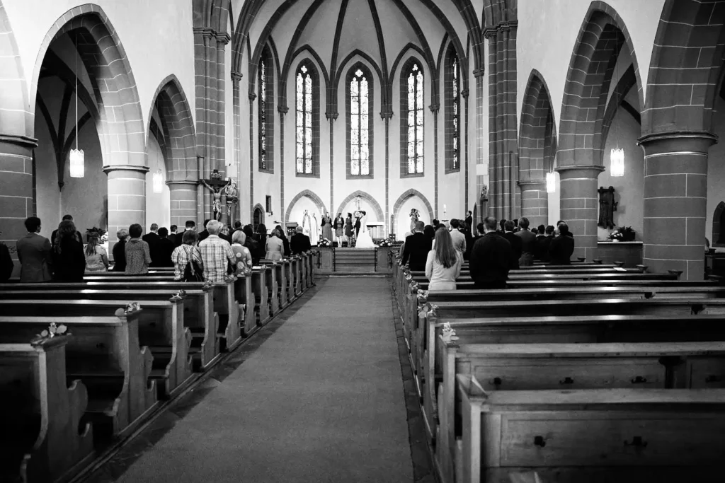 Onelovephoto Schwarz-Weiß-Foto eines Kirchenraums während einer Hochzeitszeremonie. Die Gäste sitzen in den Kirchenbänken und blicken auf den Altar, vor dem das Paar und die Trauzeugen unter hohen Bogenfenstern stehen. Hochzeitsfotografie in Mannheim und Heidelberg
