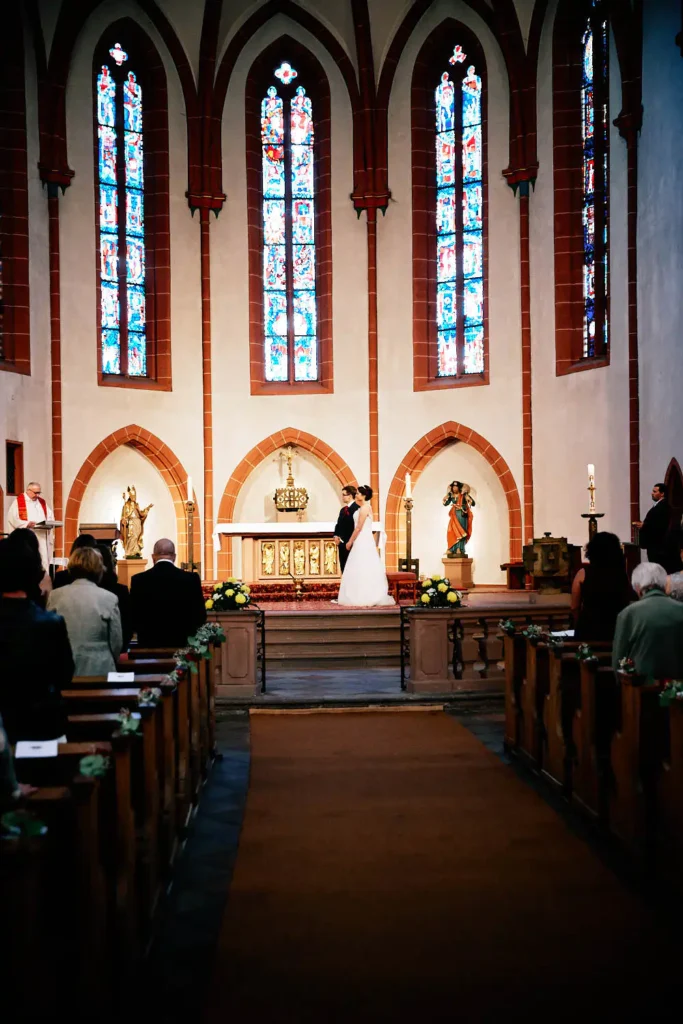 Onelovephoto Ein Brautpaar steht in einer Kirche mit hohen Glasfenstern vor dem Altar, umgeben von Gästen, die während der Hochzeitszeremonie in den Kirchenbänken sitzen. Hochzeitsfotografie in Mannheim und Heidelberg