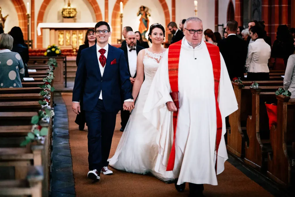 Onelovephoto Eine Braut und ein Bräutigam gehen Hand in Hand lächelnd den Gang einer Kirche entlang, wobei sie von einem Priester in weißem Gewand angeführt werden. Die Gäste schauen von den Kirchenbänken aus zu, und im Hintergrund ist der Altar zu sehen. Hochzeitsfotografie in Mannheim und Heidelberg