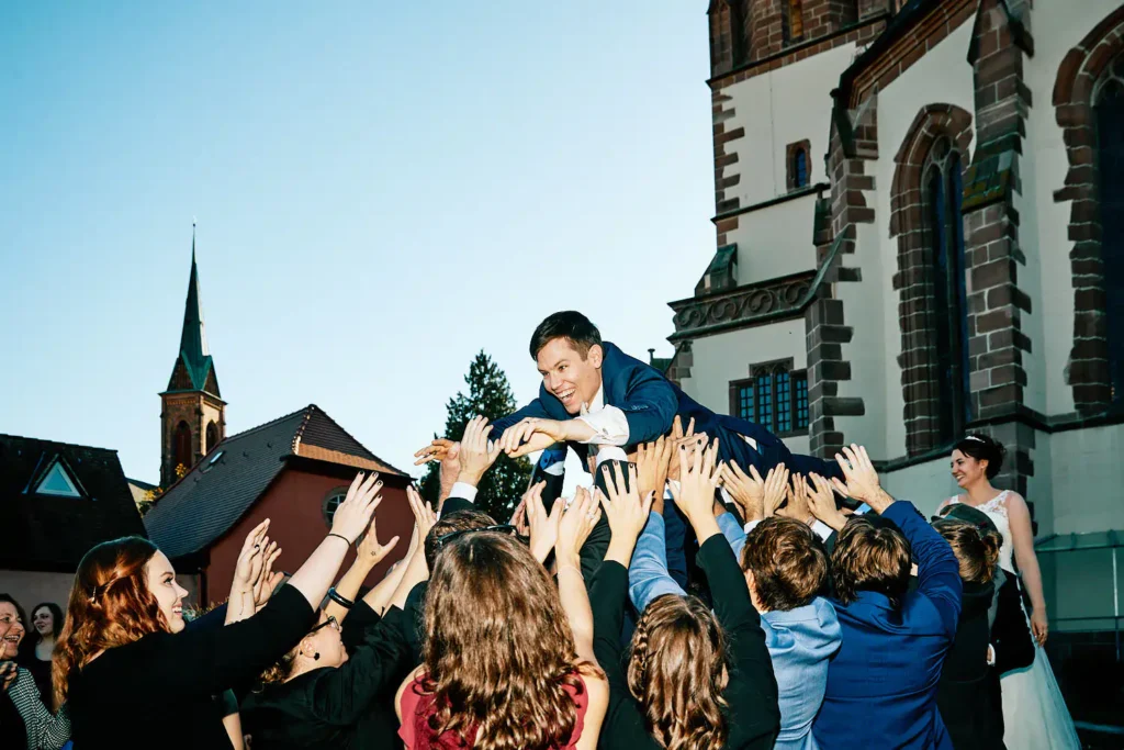 Onelovephoto Eine Gruppe von Menschen hebt vor einer Kirche freudig einen Mann im Anzug in die Luft, während eine Frau in einem weißen Kleid lächelnd zusieht. Die Szene ist festlich, mit blauem Himmel und historischen Gebäuden im Hintergrund. Hochzeitsfotografie in Mannheim und Heidelberg
