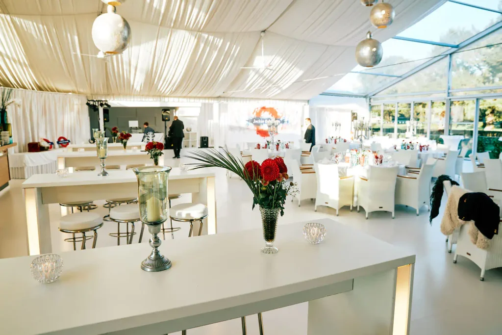 Onelovephoto Ein heller, eleganter Veranstaltungsraum mit weißen Vorhängen, modernem Dekor und Glaswänden. Die Tische sind mit roten und weißen Blumenkränzen gedeckt, und ein paar Leute bereiten den Raum vor. Hochzeitsfotografie in Mannheim und Heidelberg