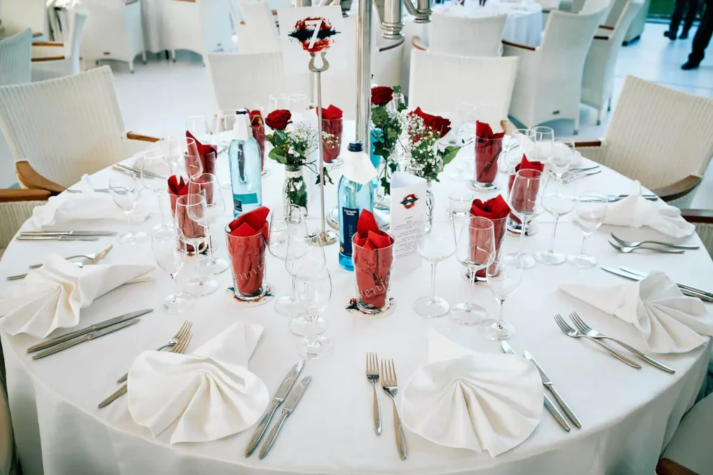 Onelovephoto Ein runder Esstisch für acht Personen, mit weißer Tischdecke, ordentlich gefalteten Servietten, roten Servietten in Gläsern, Wasser in Flaschen, Weingläsern, Besteck und kleinen Blumensträußen. Weiße Stühle umgeben den Tisch. Hochzeitsfotografie in Mannheim und Heidelberg