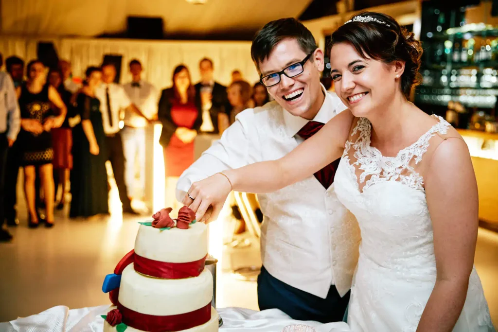 Onelovephoto Ein lächelndes Brautpaar schneidet gemeinsam die Hochzeitstorte an, umgeben von formell gekleideten Gästen bei einem hell erleuchteten Empfang. Hochzeitsfotografie in Mannheim und Heidelberg