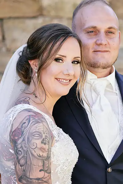 Onelovephoto Eine lächelnde Braut mit einer großen Tätowierung auf dem Arm steht neben einem Bräutigam in Anzug und Krawatte, beide posieren eng beieinander im Freien vor einer Steinmauer. Hochzeitsfotografie in Mannheim und Heidelberg Onelovephoto Eine lächelnde Braut mit einer großen Tätowierung auf dem Arm steht neben einem Bräutigam in Anzug und Krawatte, beide posieren eng beieinander im Freien vor einer Steinmauer. Hochzeitsfotografie in Mannheim und Heidelberg